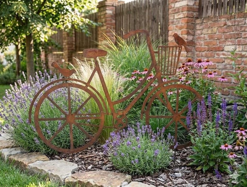 A rust-colored metal bicycle sculpture is surrounded by a lush garden filled with purple flowers and greenery.