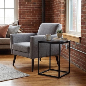 a cozy living room with a gray armchair, a black metal side table, and a small plant on the table. The room has a brick wall and a window, creating a warm and inviting atmosphere.