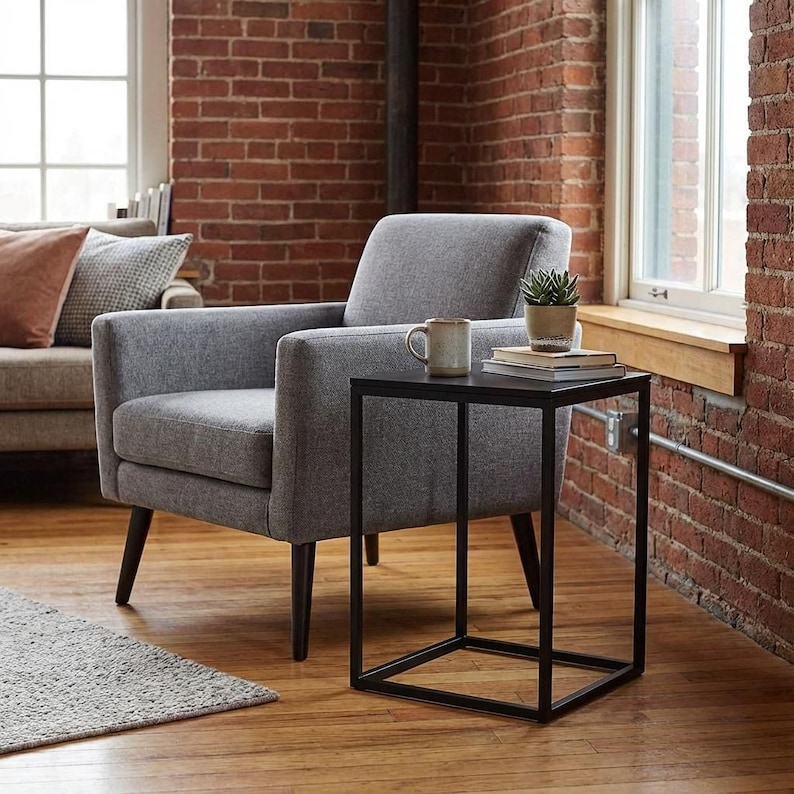 a cozy living room with a gray armchair, a black metal side table, and a small plant on the table. The room has a brick wall and a window, creating a warm and inviting atmosphere.