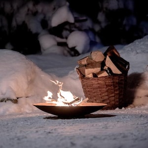 May include: A fire pit with a small fire burning inside, surrounded by snow. A wicker basket filled with firewood sits next to the fire pit.