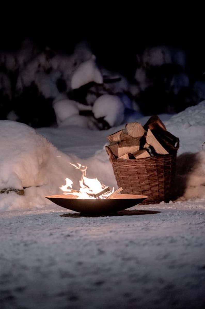 May include: A fire pit with a small fire burning inside, surrounded by snow. A wicker basket filled with firewood sits next to the fire pit.