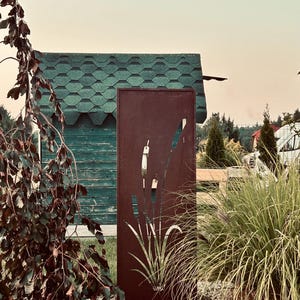 a rust-colored metal sculpture with a plant design, situated in a garden setting with a green-roofed building and lush vegetation in the background.