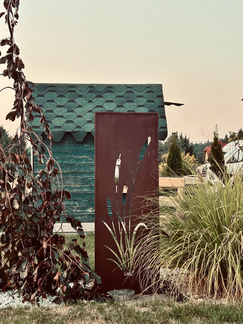 a rust-colored metal sculpture with a plant design, situated in a garden setting with a green-roofed building and lush vegetation in the background.