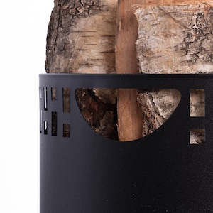 May include: A black metal log holder with decorative cutouts, filled with birch and other wood logs. The logs have a natural, textured appearance, with varying shades of brown and tan. The holder is cylindrical and sits on a white background.