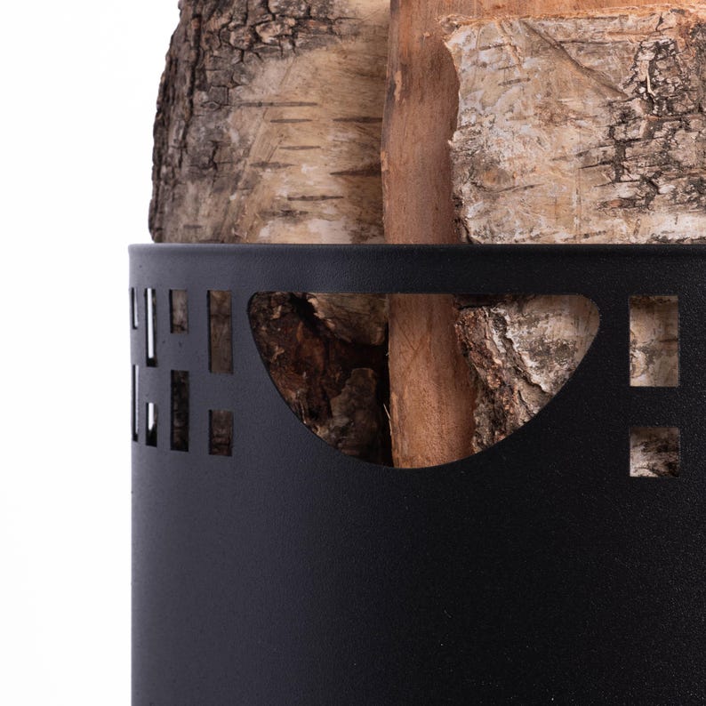 May include: A black metal log holder with decorative cutouts, filled with birch and other wood logs. The logs have a natural, textured appearance, with varying shades of brown and tan. The holder is cylindrical and sits on a white background.