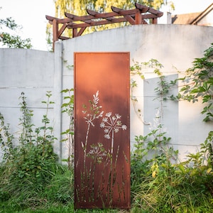 a rust-colored metal gate with a white floral design on it, standing in a garden setting with a white wall and greenery in the background.