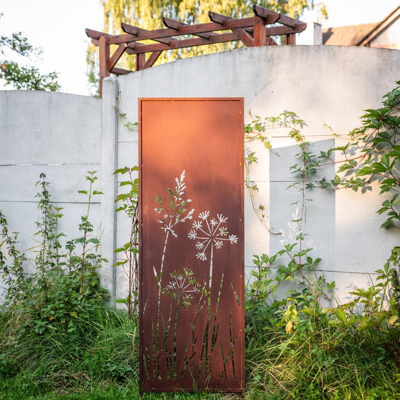 a rust-colored metal gate with a white floral design on it, standing in a garden setting with a white wall and greenery in the background.
