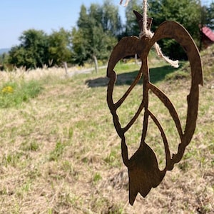 Op de afbeelding: Een roestig metalen bladvormig ornament dat aan een touwtje hangt. Het ornament heeft de vorm van een blad met een gedetailleerd adernetwerk. Het ornament hangt voor een grasveld en bomen.