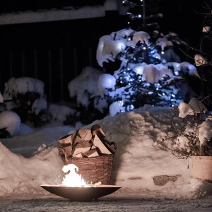 May include: A fire pit with a small fire burning inside, surrounded by snow. A basket of firewood sits nearby, and a Christmas tree with white lights is in the background.
