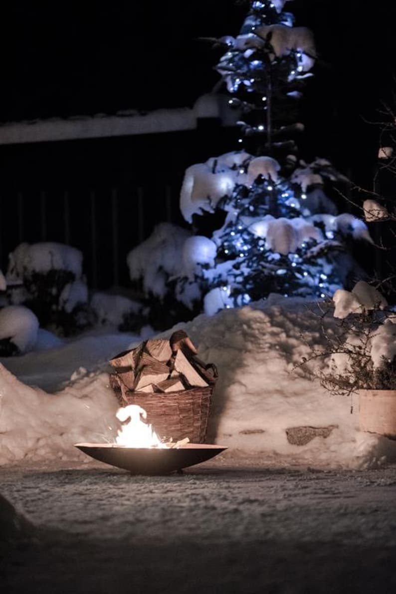 May include: A fire pit with a small fire burning inside, surrounded by snow. A basket of firewood sits nearby, and a Christmas tree with white lights is in the background.