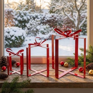 Op de afbeelding: Drie rode metalen cadeauverpakkingen met rode strikken. De dozen zijn verschillend van grootte en staan op een houten oppervlak met kerstversieringen en dennenappels. Door het raam is een besneeuwde winterscène te zien.