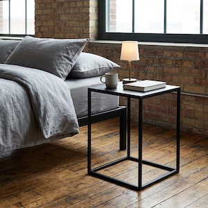 a cozy bedroom scene with a bed, a black metal side table, and a lamp on the table. The bed is covered in a gray comforter and has two pillows. The side table has a lamp and a book on it. The room has a brick wall and a window, creating a warm and inviting atmosphere.