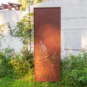 Könnte beinhalten: Ein hoher, rechteckiger, rostbrauner Metall-Gartenzaun mit ausgeschnittenen Farn- und Pflanzenmotiven. Der Zaun steht in einer grasbewachsenen Fläche mit einer Betonmauer und Grün in der Hintergrund. Er wird von zwei dünnen Metallbeinen getragen.