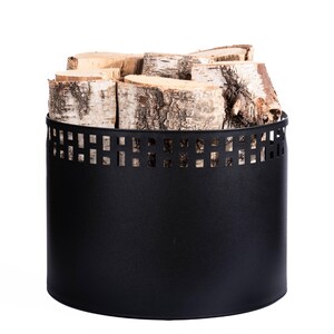 May include: A black metal log holder filled with birch firewood. The cylindrical holder has a decorative square cut-out pattern near the top. The birch logs are light brown and white, with visible bark.