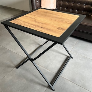 Könnte beinhalten: Ein quadratischer Beistelltisch mit einer Holzmaserungsplatte und einem schwarzen Metallrahmen. Der Tisch hat einen schwarzen Rand um die Tischplatte. Die Beine sind X-förmig. Der Tisch ist etwa 61 cm groß.
