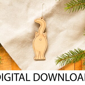 May include: A wooden cat ornament with a heart on its backside. The ornament is light brown and has a string for hanging. The ornament is on a white linen surface with a green pine branch.