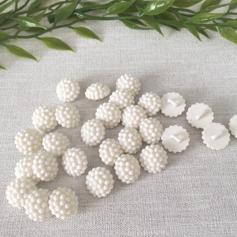 May include: A collection of 20 white plastic buttons with a textured, raised dot design. The buttons are arranged in a semi-circle on a white surface.