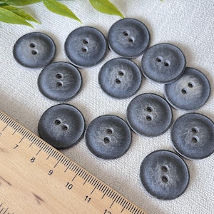 May include: A collection of 12 grey buttons with two holes each. The buttons are made of a textured material and have a slightly distressed look.