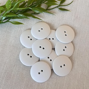 May include: A cluster of white, circular buttons, each with two holes. The buttons are piled together on a light-coloured surface, with green leaves in the background. These buttons are suitable for sewing and craft projects.