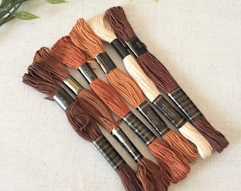 Puede incluir: Seis carretes de hilo de bordar en tonos marrones, naranjas y beige. Los carretes están etiquetados con el nombre de la marca "Duchess" y los números de color "220", "927", "812" y "810".
