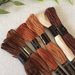 Puede incluir: Seis carretes de hilo de bordar en tonos marrones, naranjas y beige. Los carretes est&aacute;n etiquetados con el n&uacute;mero de color y el nombre de la marca "DMC" y "Duchess".