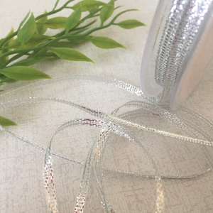 May include: Silver ribbon with a metallic sheen, wound around a spool. The ribbon is thin and delicate, with a subtle shimmer.