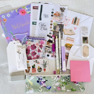 May include: A stationery set featuring a purple floral notebook, a spiral-bound floral notepad, various sticker sheets with botanical and book-themed illustrations, a pink sticky note pad, three pens, a highlighter, and a floral print pencil case.