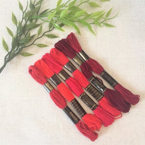 May include: Six spools of embroidery floss in shades of red and burgundy. The floss is wrapped around cardboard spools with gold labels that include the brand name "Anchor" and the text "6 Strands Embroidery Thread 100% Cotton".