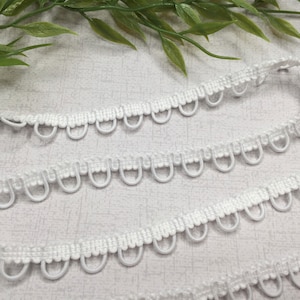 May include: White looped trim with a scalloped edge. The trim is made of a woven material and features small, evenly spaced loops. The trim is shown against a light background with green foliage in the upper left corner.