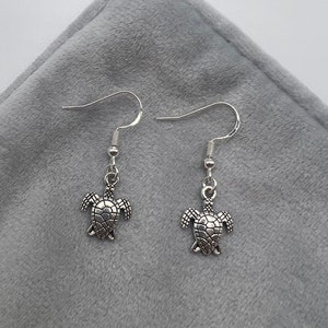 May include: A pair of silver dangle earrings featuring a turtle charm. The turtle has a detailed shell and is hanging from a silver hook.