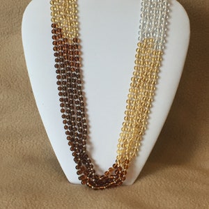 May include: A multi-strand necklace on a white display bust. The necklace has strands of small, round beads in shades of brown, amber, and clear. The beads are strung together, creating a layered effect with a gradient of colours.