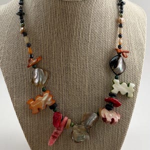 May include: A colorful necklace with a variety of beads and shell-shaped pendants. The necklace features black, orange, red, and iridescent shell elements, strung together with smaller beads. It is displayed on a neutral-toned mannequin.