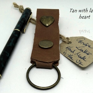 May include: Tan leather keyring with a large heart-shaped metal charm and a smaller round metal charm. The keyring also has a metal ring. The tag reads "Genuine Leather Hand Made in the UK".