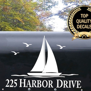 May include: A white sailboat with a white wave design on a black mailbox with the address 225 Harbor Drive. The mailbox is outdoors with green trees in the background.