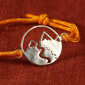 Op de afbeelding: Een zilveren armband met een bedel in de vorm van een bergketen. De armband is gemaakt van een geel koord.