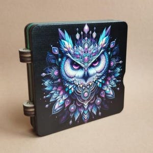 Puede incluir: Una caja de madera negra con un lado con bisagras. La caja presenta una ilustración detallada de un búho con plumas azules, moradas y blancas. El búho tiene ojos grandes e intensos y una corona decorativa.