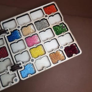 K&ouml;nnte beinhalten: Ein Holzperlen-Organizer mit mehreren F&auml;chern, die mit bunten Perlen gef&uuml;llt sind. Die Perlen sind in verschiedenen Farben erh&auml;ltlich, darunter Rot, Orange, Gelb, Rosa, Blau, Gr&uuml;n und Silber. Der Organizer ist f&uuml;r kleine Bastelmaterialien konzipiert.