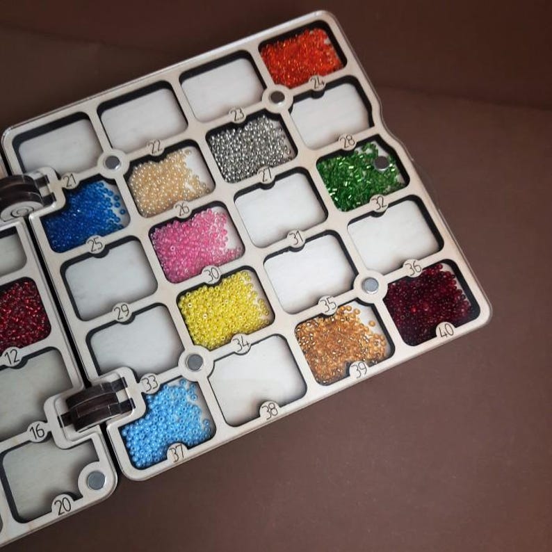 K&ouml;nnte beinhalten: Ein Holzperlen-Organizer mit mehreren F&auml;chern, die mit bunten Perlen gef&uuml;llt sind. Die Perlen sind in verschiedenen Farben erh&auml;ltlich, darunter Rot, Orange, Gelb, Rosa, Blau, Gr&uuml;n und Silber. Der Organizer ist f&uuml;r kleine Bastelmaterialien konzipiert.