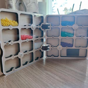 May include: A two-part wooden organiser with clear compartments filled with colourful beads. The organiser is hinged and has a light-coloured frame with a black border. The compartments are numbered for easy organisation.