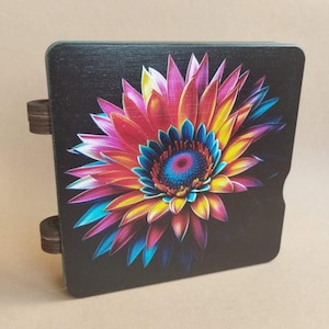 Puede incluir: Una caja de madera con un diseño floral vibrante y colorido. La flor presenta tonos de rojo, rosa, amarillo, azul y morado sobre un fondo negro. La caja tiene dos bisagras de madera en el lateral.