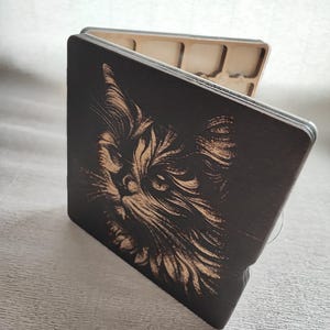 K&ouml;nnte beinhalten: Eine Holzbox mit einer detaillierten Gravur eines Katzenkopfes. Das Fell der Katze ist aufwendig in das dunkle Holz ge&auml;tzt, was einen auff&auml;lligen Kontrast erzeugt. Die Box ist quadratisch und hat einen Klappdeckel mit hellem Innenraum.