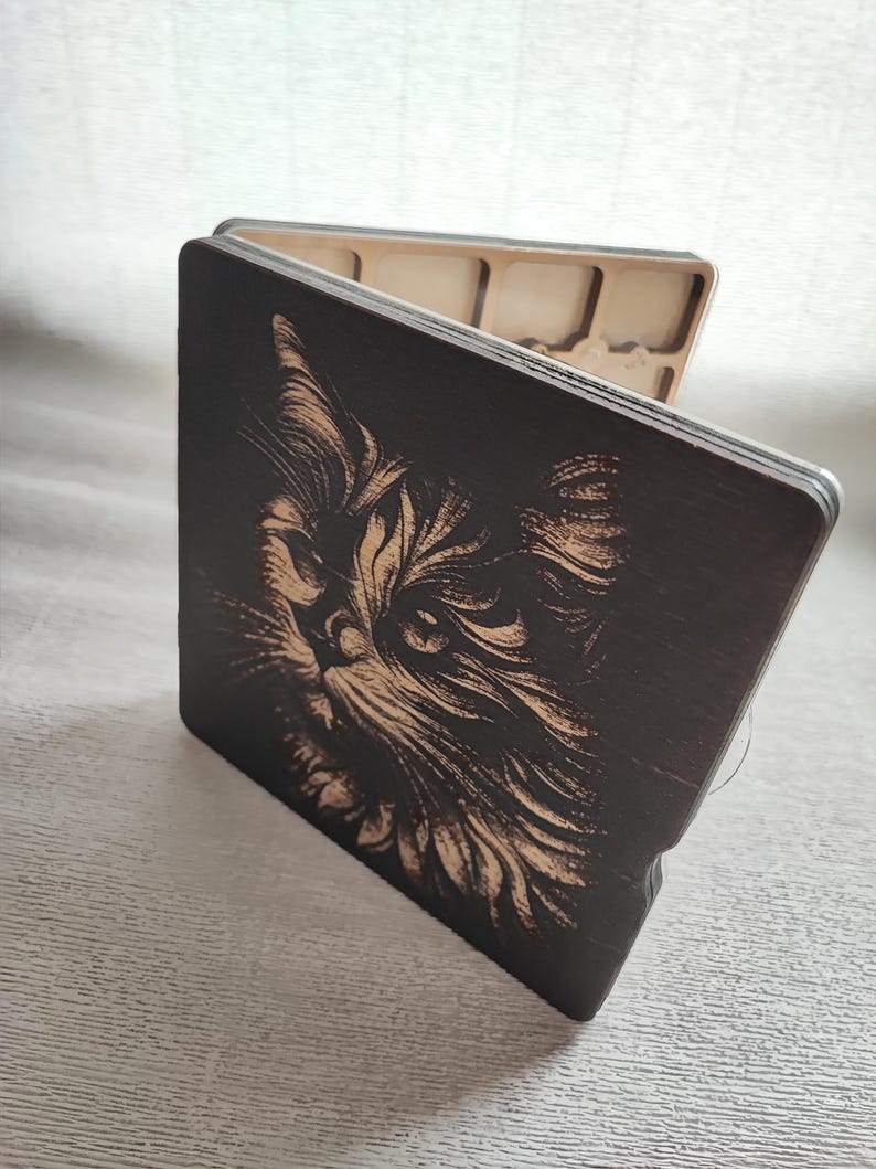 K&ouml;nnte beinhalten: Eine Holzbox mit einer detaillierten Gravur eines Katzenkopfes. Das Fell der Katze ist aufwendig in das dunkle Holz ge&auml;tzt, was einen auff&auml;lligen Kontrast erzeugt. Die Box ist quadratisch und hat einen Klappdeckel mit hellem Innenraum.