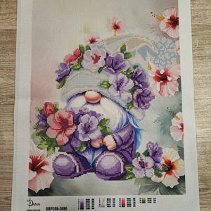 Puede incluir: Un kit de punto de cruz con un gnomo sosteniendo flores, rodeado de hibiscos. El gnomo viste un atuendo morado y un sombrero adornado con flores. El kit incluye el texto "Stitch Bloom" y el código de producto SBP359-3695.