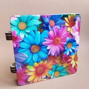 Puede incluir: Caja decorativa cuadrada con un vibrante diseño floral. Las flores son en tonos de azul, rosa, amarillo y naranja, con una apariencia detallada y realista. La caja tiene dos pequeños pomos cilíndricos de madera en el lateral.