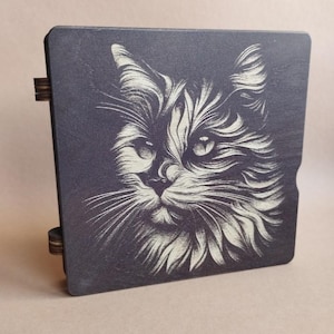 K&ouml;nnte beinhalten: Eine dunkelgraue Holzkiste mit einer detaillierten Illustration eines Katzenkopfes. Das Fell der Katze ist in Wei&szlig; dargestellt und bildet einen auff&auml;lligen Kontrast zum dunklen Hintergrund. Die Kiste hat Holzscharniere.