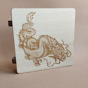 K&ouml;nnte beinhalten: Eine hellfarbene Holzkiste mit einer detaillierten Gravur eines Drachen. Der Drache ist aufwendig geschnitzt und zeigt Schuppen, Krallen und eine flie&szlig;ende M&auml;hne. Die Kiste hat dunkelbraune Scharniere.