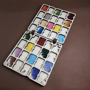 K&ouml;nnte beinhalten: Ein h&ouml;lzerner Perlenorganizer mit zwei Scharnierabschnitten, die jeweils nummerierte F&auml;cher mit farbigen Perlen enthalten. Die Perlen sind in verschiedenen Farben, darunter Rot, Gelb, Gr&uuml;n, Blau und Rosa. Der Organizer ist ge&ouml;ffnet.