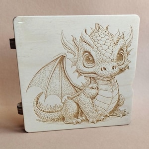 K&ouml;nnte beinhalten: Eine hellfarbene Holzkiste mit einem detaillierten gravierten Bild eines Babydrachens. Der Drache hat gro&szlig;e Augen, Fl&uuml;gel und ein detailliertes Schuppenmuster. Die Box hat dunkle Scharniere an der Seite.