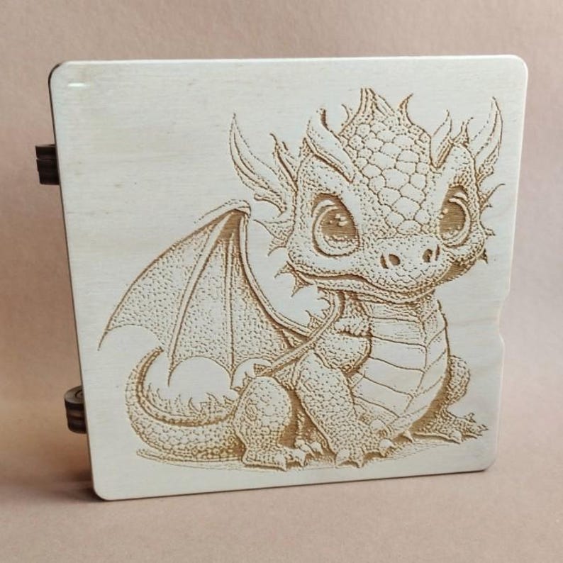 K&ouml;nnte beinhalten: Eine hellfarbene Holzkiste mit einem detaillierten gravierten Bild eines Babydrachens. Der Drache hat gro&szlig;e Augen, Fl&uuml;gel und ein detailliertes Schuppenmuster. Die Box hat dunkle Scharniere an der Seite.