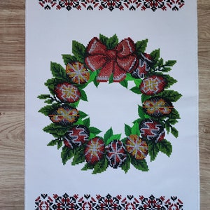May include: White embroidered kitchen towel featuring a red, green and yellow floral wreath design. The wreath is made of stylised flowers and leaves. The towel has a white background with a red and black border.